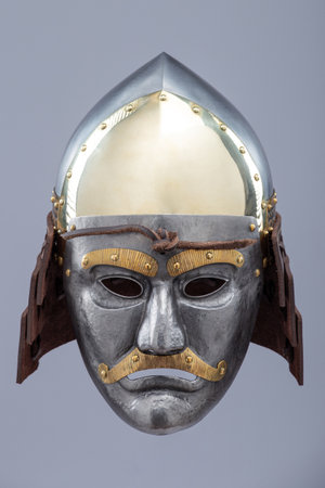 Historical metallic combat helmet with integrated face mask. Pointed steel helmet featuring a detailed metallic face mask with eye openings, decorative elements, and side protection flaps.の写真素材