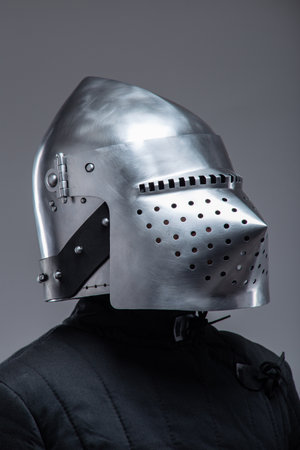 Close-Up of a Medieval Knight's Helmet with Perforated Visor. This image focuses on a shiny metal helmet with numerous small holes in the visor, worn by a person in a dark quilted garment.の写真素材