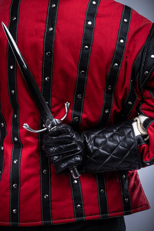 Person Holding a Sharp Blade Behind Their Back. This image shows a person wearing a red and black padded jacket and black gloves, holding a silver blade with a complex hilt behind their back.の写真素材