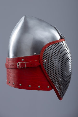 Modern fencing helmet with protective mesh visor. A shiny silver helmet features a full face mesh visor and is secured with a red leather strap and metal buckle.の写真素材