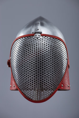 Modern fencing helmet with protective mesh visor. A shiny silver helmet features a full face mesh visor and is secured with a red leather strap and metal buckle.の写真素材