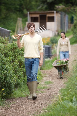 Man holding spading fork, woman pushing wheelbarrow with potted flowers in the backgroundの写真素材