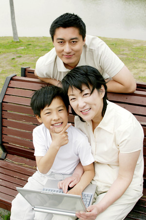 A woman and her son sitting on the bench with a laptop on their laps while her husband standing behind themの写真素材