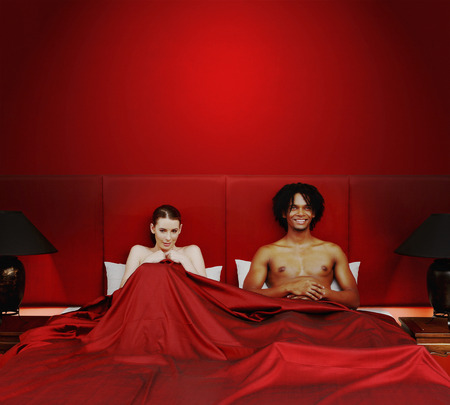 A shy naked lady and African American man sitting on the bed covered with blanketの写真素材