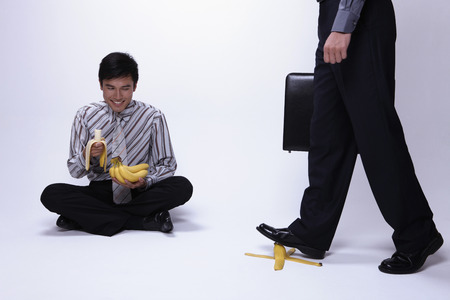 Businessman sitting on the floor eating banana while watching another businessman about to step on banana peelの写真素材
