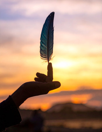 Feather in hand on sunset background. Selective focus. Shallow depth of fieldの素材