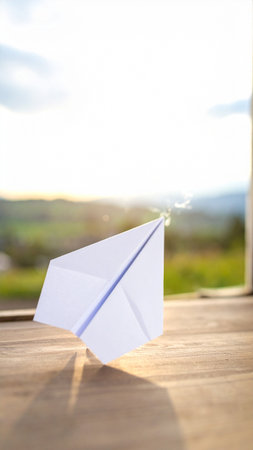 Paper airplane on a wooden table in front of a window at sunsetの素材