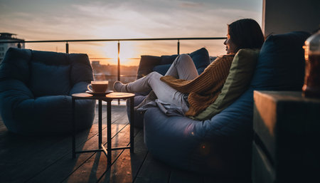 Beautiful young woman relaxing on a balcony at sunset, drinking coffee and watching the sunsetの素材