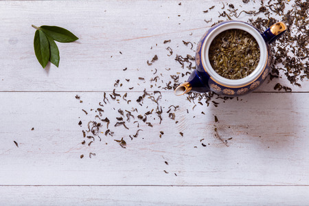 Teapot and dry tea leaves on white wooden background, green tea leaf on table, black herbal homemade hot beverage in porcelain pot, english tea ceremony background, top view from above, copy spaceの写真素材