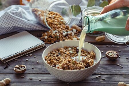 Female hand holding glass bottle pouring milk in cereal granola flakes bowl with nuts seeds raisins on brown wooden table background, healthy breakfast lifestyle concept, home muesli food oat mealの写真素材