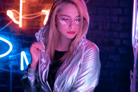 Beautiful young woman wearing metallic bomber jacket trendy stylish glasses stand near brick wall illuminated with neon lights, pretty fashion girl in modern cloth eyewear in night glow on nightclubの写真素材
