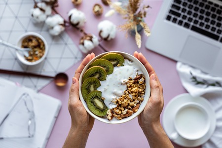 Female hand holding healthy breakfast bowl over work table background concept enjoy detox morning meal with laptop milk, woman eat natural granola nutrition detox food in home office, top viewの写真素材