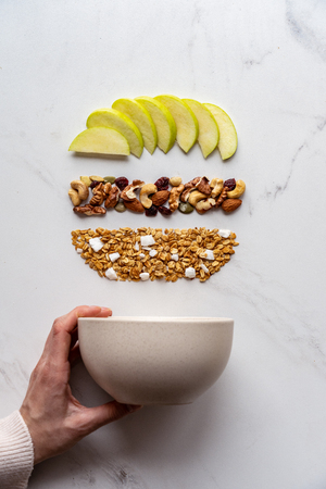 Healthy breakfast concept, female hand holding bowl with granola nuts apple on white background, morning organic food with fruit diet on table, vegan detox meal for health nutrition vertical top viewの写真素材