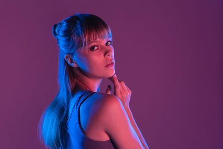 Fashion attractive young blond hair 20s fashion girl model pretty face looking at camera standing at purple studio background, sexy trendy stylish woman in pink neon party night glow concept portraitの写真素材
