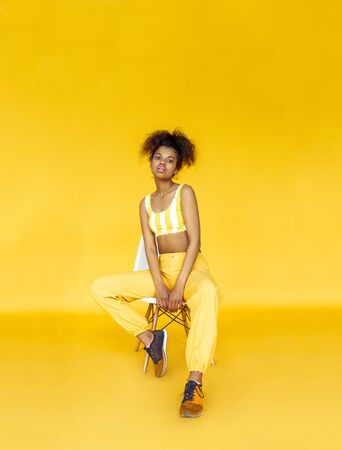 Fashion young african girl black woman wear stylish yellow clothes looking at camera sit on chair isolated on studio wall background, vertical banner for website design, portrait, copy spaceの写真素材