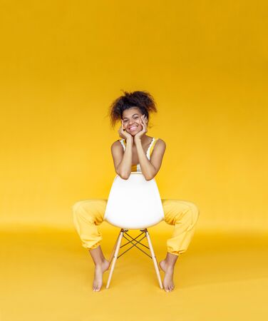 Smiling young african woman wear stylish yellow clothes sit on chair isolated on background, happy black teen fashion girl looking at camera on summer studio wall, vertical banner design, copy spaceの写真素材