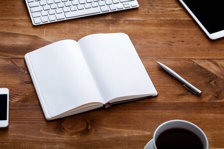 Work space desk with open paper empty white blank page notebook for notes pen and devices on brown...