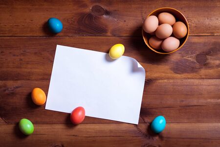 Happy easter background, multicolored eggs and blank white paper on brown wooden table board, spring fun decoration design, traditional april celebration greeting card on decorated wood desk backdropの写真素材