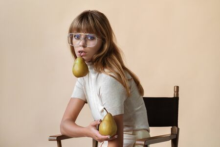 Fashion young lady sit on chair hold pears look at camera on beige.の写真素材