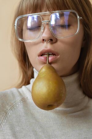 Fashion young lady hold pear in mouth on beige background, vertical.の写真素材