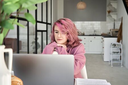Young hipster girl with pink hair looking at laptop sit at kitchen table.