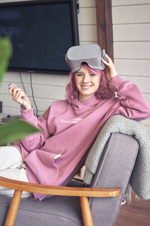 Happy hipster teen girl wear vr headset hold controller look at camera in chair.