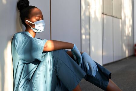 Tired exhausted woman african nurse wear face mask gloves sit on hospital floor.の写真素材