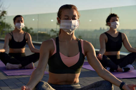 Fit sporty young woman wear face mask meditating keep social distance at group class.
