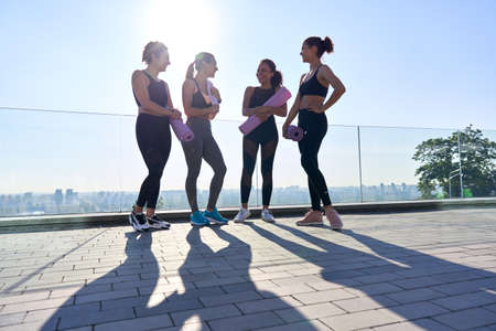 Happy diverse fit girls friends laugh stand hold mats group yoga class outdoor.