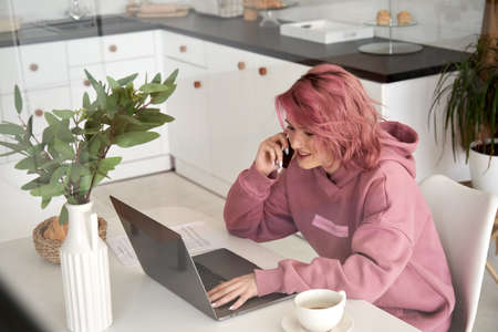 Smiling hipster teen girl talking on phone using laptop sitting at home office table.の写真素材