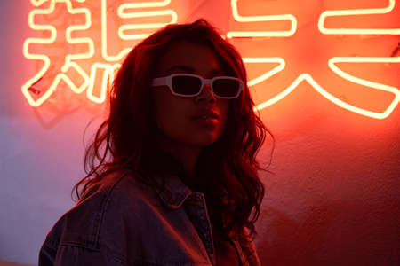 African woman wears trendy glasses stands in red neon backlight in night club.