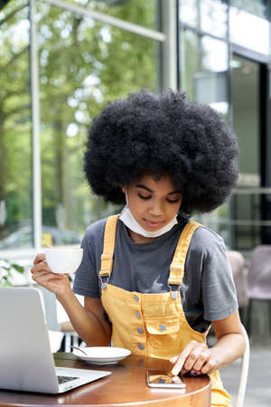African teen girl wearing face mask using phone sit at table in outdoor cafe.の写真素材