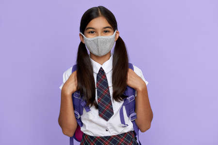 Indian school girl wearing face mask standing on lilac background, portrait.の写真素材