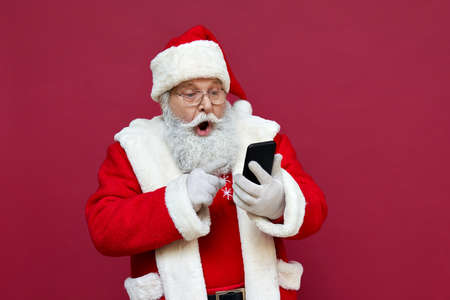 Surprised Santa Claus using smart phone isolated on red background.