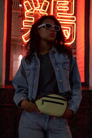 Cool African hipster lady wears trendy glasses stands under neon night sign.