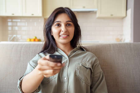 Happy indian woman sitting on sofa holding remote control watching tv at home.