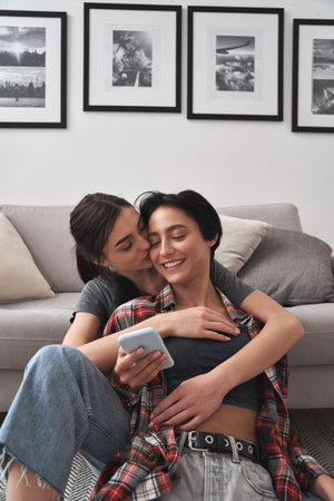 Young happy lesbian couple hugging using phone together at home.の写真素材