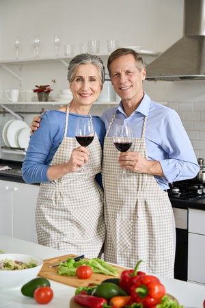 Happy old couple in aprons hugging, drinking wine, looking at camera in kitchen.の写真素材