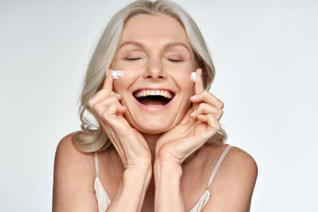 Happy funny 50s middle aged woman applying cream on face isolated on white.