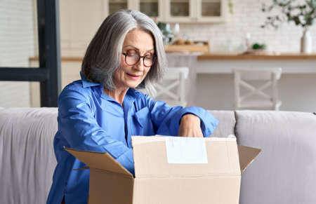 Senior mid age 60s woman opening parcel box at home on the couch.