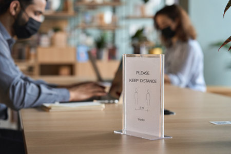 Closeup of social distance sign standing on table with people working on pc.