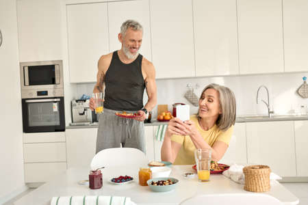 Happy older mature family couple having breakfast using phone in kitchen.