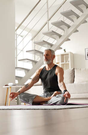 Healthy senior middle aged tattooed man meditating in yoga pose at home.