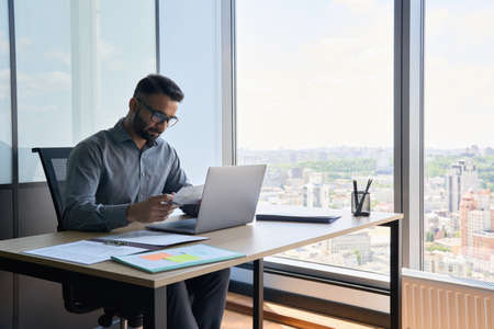 Serious indian financial analyst working on document using laptop in office.の写真素材