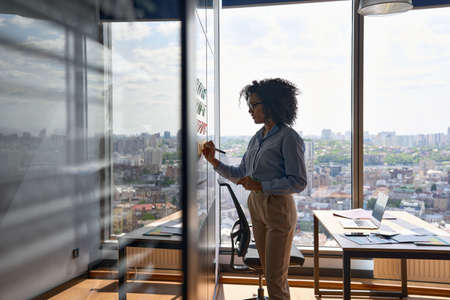Afro American ceo writing on sticky notes in office with big city urban view.