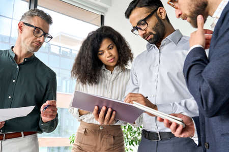Diverse executive team people discussing project report standing in boardroom.
