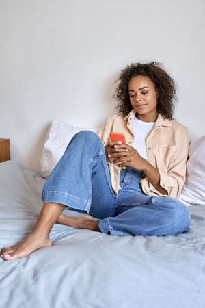 Young happy afro American mixed race girl using cell phone sit on bed relaxed.の写真素材