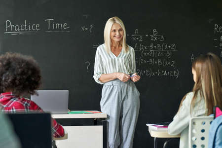 Smiling young teacher having mathematics lesson standing at chalkboard.