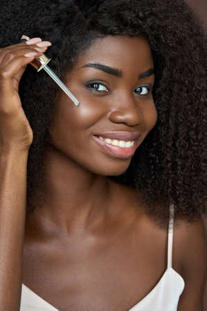 Groomed happy black girl applying pipette facial serum oil. Skincare concept.
