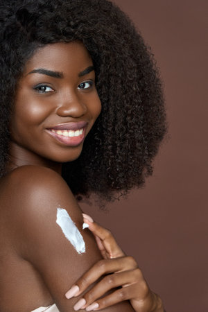 Smiling happy young afro american girl applying cream on body. Skincare concept.の写真素材